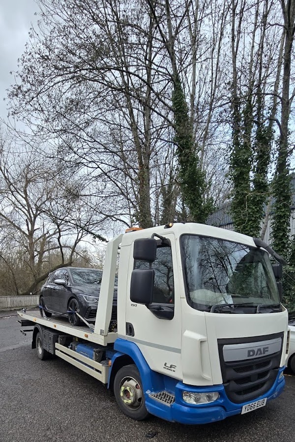Prompt Recovery DAF LF flatbed truck transporting a dark saloon car, parked on a road with bare winter trees in the background