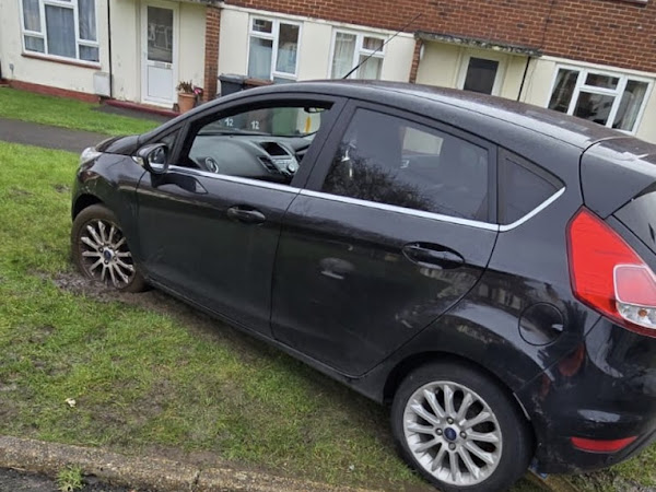 Black Ford Fiesta hatchback broken down and tilted on the grass verge outside a residential block of flats, awaiting recovery with Prompt Recovery