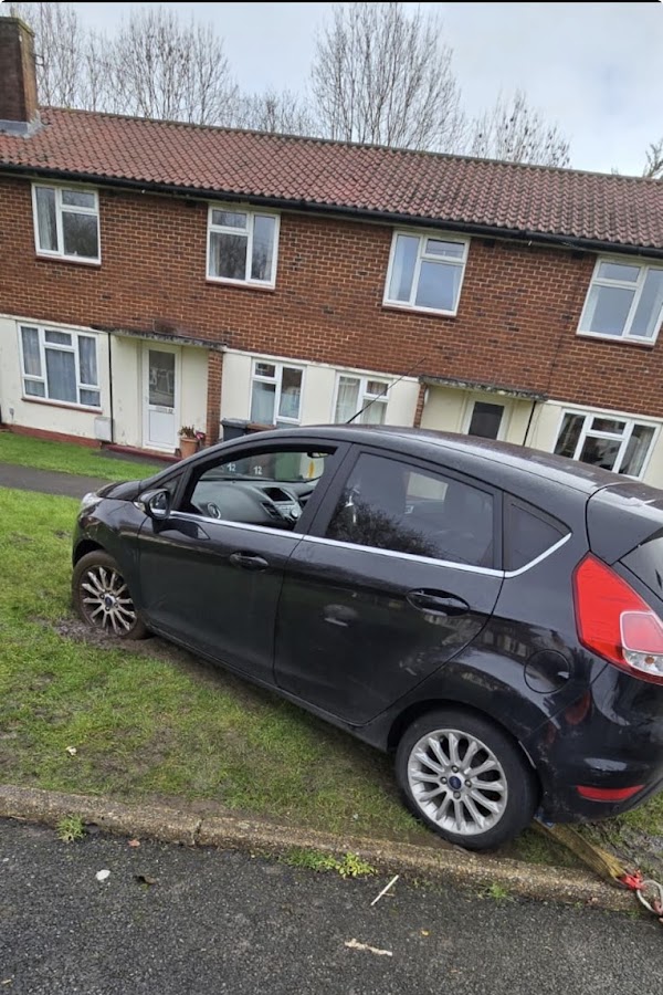 Black Ford Fiesta hatchback broken down and tilted on the grass verge outside a residential block of flats, awaiting recovery with Prompt Recovery