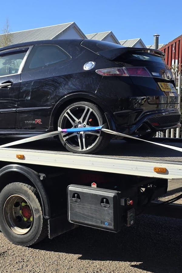 Black Honda Civic Type R sports car secured with straps on a Prompt Recovery DAF LF flatbed truck in a yard on a sunny day
