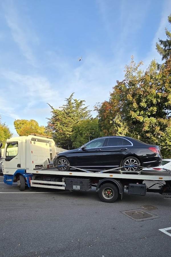 Prompt Recovery DAF LF flatbed truck transporting a black Mercedes C-Class saloon in a car park surrounded by autumn trees