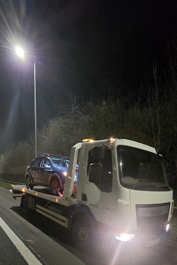 Prompt Recovery DAF LF flatbed truck at night on a roadside with a dark crossover SUV secured on the bed, amber warning lights on