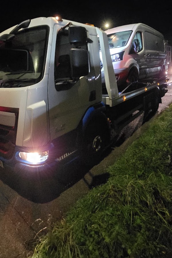 Prompt Recovery DAF LF flatbed truck at night on a grass verge recovering a large white van, headlights illuminating the dark roadside