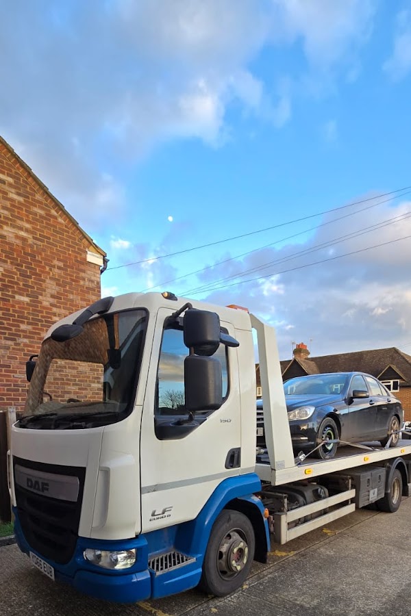 Prompt Recovery DAF LF flatbed truck transporting a dark BMW saloon on a residential road at dusk