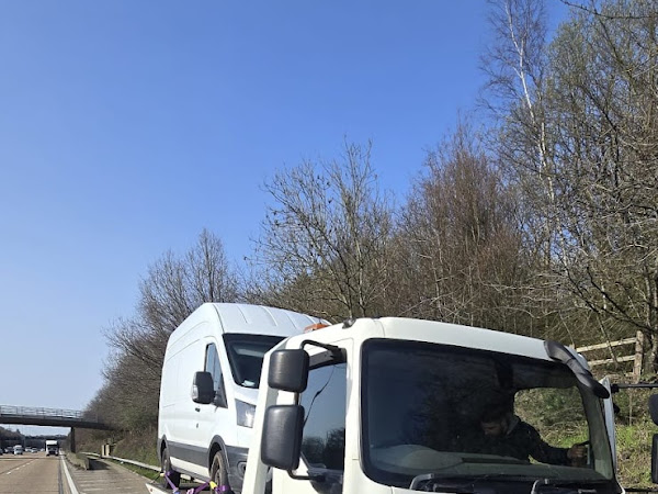 Prompt Recovery DAF LF flatbed truck on a motorway hard shoulder recovering a white panel van under a clear blue sky