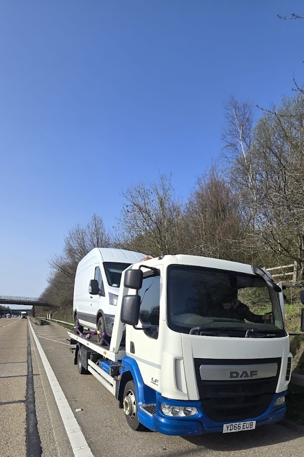 Prompt Recovery DAF LF flatbed truck on a motorway hard shoulder recovering a white panel van under a clear blue sky