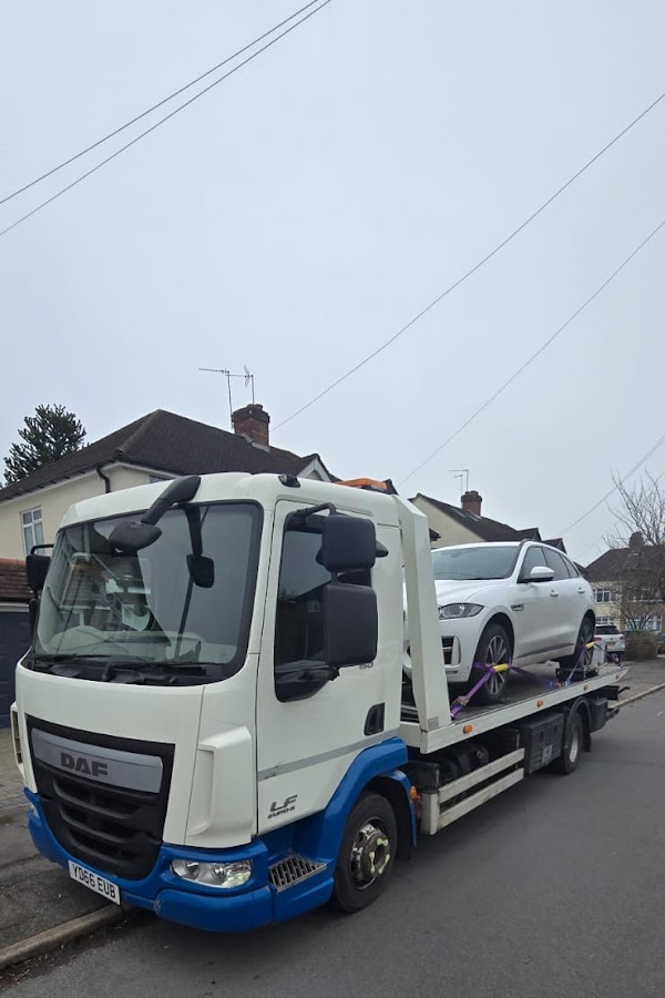 Prompt Recovery DAF LF flatbed truck with a white Jaguar F-Pace SUV secured on the bed, collected from a residential street on an overcast day