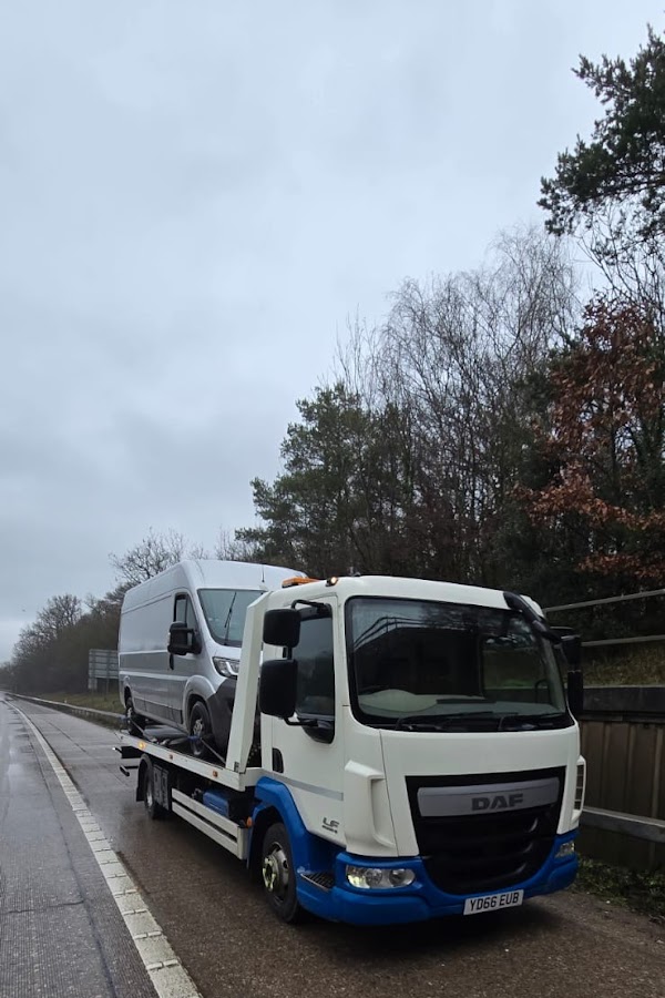 Prompt Recovery DAF LF flatbed truck on a wet dual carriageway recovering a large white Mercedes Sprinter van in overcast conditions